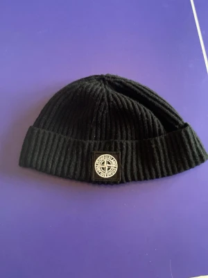 Svart ribbad mössa Stone Island - Säljer en svart ribbstickad mössa från Stone Island med uppvikt kant och den klassiska logotypen framtill. Mössan är i mjukt material och har en clean, stilren look som passar perfekt till streetwear och vardagsoutfits.