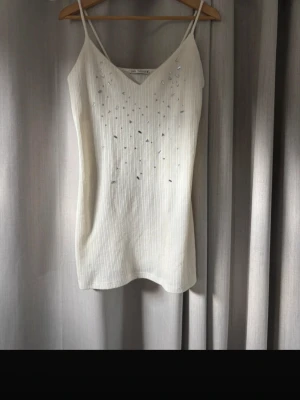 Vit ribbad klänning med paljetter Zara - Säljer en vit ribbad kortklänning från Zara med tunna axelband och v-ringning. Klänningen har dekorativa paljetter i olika former framtill som ger en unik touch. Perfekt för dig som gillar stilrena men ändå detaljerade plagg.