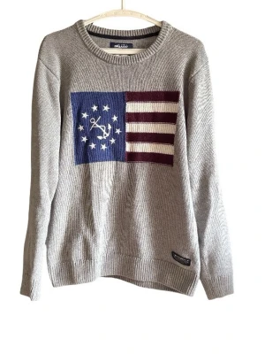 Grå stickad tröja med flaggmönster - Grå stickad tröja från Sebago med ett stort flagginspirerat mönster framtill i blått, vinrött och vitt. Tröjan har rund halsringning, långa ärmar och ribbade muddar. Perfekt för dig som gillar klassisk och nautisk stil.