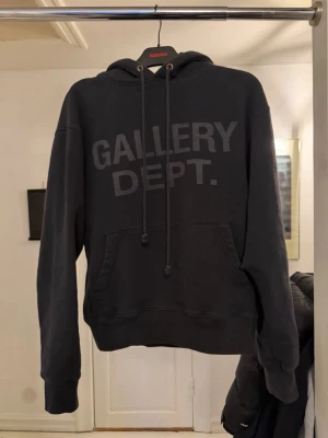 Svart hoodie från Gallery Dept. - Svart hoodie från Gallery Dept. med stor logga tryckt på bröstet. Klassisk känguruficka framtill och justerbar dragsko i huvan. Mjuk bomullskänsla och avslappnad passform. Har ett litet hål som blivit ihopsydd på ena armen. Inte märkbart 