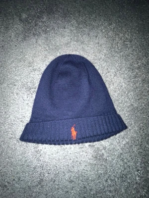 Mörkblå mössa från Polo Ralph Lauren - Mörkblå stickad mössa från Polo Ralph Lauren med klassisk orange broderad logga framtill. Mössan har uppvikt kant och är tillverkad i mjukt material som passar perfekt för kyliga dagar.
