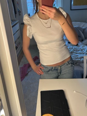 Vit ribbad topp med volangärmar - Snygg vit ribbad topp med korta volangärmar och rund halsringning. Toppen har en figurnära passform och är tillverkad i ett mjukt bomullsmaterial. Perfekt för dig som vill ha en söt och trendig look. Helt oanvänd