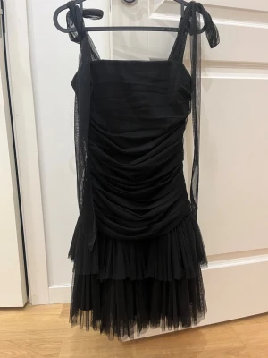 Svart kort klänning med volang - Supersnygg svart kort klänning med tunna axelband som knyts i rosetter. Klänningen har rynkade detaljer framtill och volang i nederkant för extra volym. Tillverkad i ett tunt, skirt material som ger en elegant look.