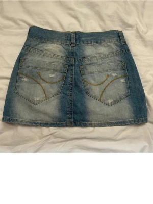 Blå jeanskjol - Snygg blå jeanskjol i kort modell med slitningar och två bakfickor med dekorativa sömmar. Kjolen har klassisk femficksdesign och stängs med knapp och dragkedja framtill. Perfekt till sommarn😍