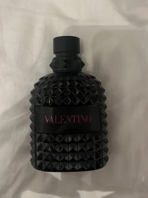 Valentino svart parfymflaska - Snygg parfym från Valentino i en svart, robust flaska med nitar och strukturmönster. Flaskan har en modern och edgy design med Valentino-loggan i rosa på svart etikett. Perfekt för dig som gillar exklusiva och stilrena dofter.