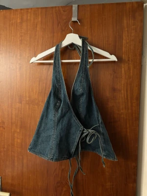 Jeansblå omlottopp med knytband - Säljer en unik jeansblå omlottopp i denim med knytband både i nacken och vid midjan. Köpt på Glassons i Australien Toppen är ärmlös och har en djup v-ringning framtill. Perfekt för dig som vill ha en cool och trendig look!