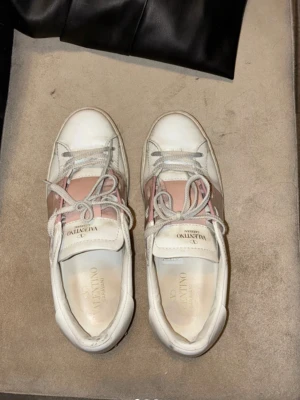 Valentino Garavani vita sneakers - Snygga vita sneakers från Valentino Garavani med rosa och beige detaljer samt klassiska nitar på hälen. Skorna har rund tå, vita snören och är tillverkade i läder. Perfekta för dig som vill ha en exklusiv touch på din outfit.Dessa Valentino Garavani är köpta från Vestiaire Collective och verifierade som äkta genom deras professionella äkthetskontroll. Kvitto saknas men äkthet är bekräftad.