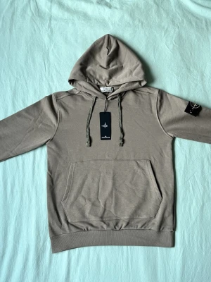Beige hoodie från Stone Island - Snygg beige hoodie från Stone Island med klassisk huva och känguruficka framtill. Ikonisk svart och gul logotyp-patch på vänster ärm. Tillverkad i mjukt bomullsmaterial och har ribbade muddar vid ärmslut och nederkant. Perfekt för en avslappnad streetwear-look.