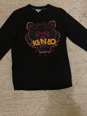 Svart Kenzo sweatshirt med tiger - Svart sweatshirt från Kenzo med ikoniskt broderat tigerhuvud i flera färger och texten 'KENZO PARIS' på bröstet. Tröjan har rund halsringning, långa ärmar och är gjord i mjukt bomullsmaterial. Perfekt statement-plagg för dig som gillar streetwear.
