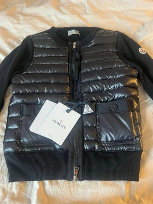 Svart dunjacka från Moncler - Snygg svart dunjacka från Moncler med quiltad front och två stora fickor med knappar. Jackan har ribbade muddar, dragkedja framtill och Moncler-logga på ärmen. Materialet är glansigt på framsidan och ärmarna är i mjukare tyg. Perfekt för kalla vinterdagar.