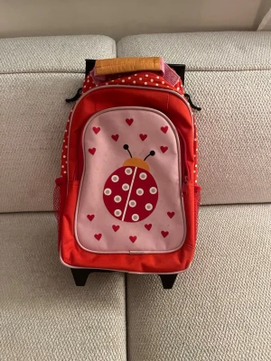 Röd barnväska med nyckelpigemotiv - Säljer en röd och rosa barnväska med draghandtag och hjul. Väskan har ett gulligt nyckelpigemotiv på framsidan och små röda hjärtan. Sidorna har vita prickar på röd botten och nätfickor. Perfekt för resor eller skola!                                VET EJ VAD DET ÄR FÖR Varumärke 