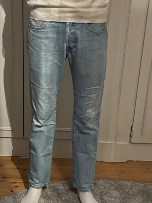 Levi's 501 ljusblå raka jeans - Säljer ett par klassiska Levi's 501 jeans i ljusblå denim. Jeansen har rak passform, fem fickor och knappgylf. Perfekta för en avslappnad och tidlös stil. Ikonisk läderpatch bak i midjan och röd Levi's-tag på bakfickan.