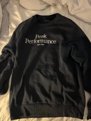 Blå Peak Performance sweatshirt - Snygg blå sweatshirt från Peak Performance med vit logga och text på bröstet. Klassisk rund halsringning och ribbade muddar vid ärmslut och nederkant. 