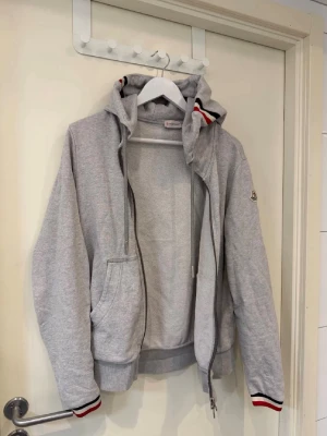 Grå hoodie från Moncler med dragkedja - Snygg grå hoodie från Moncler med dragkedja, huva och fickor framtill. Röd, vit och svart rand på muddar och huva ger en sportig vibe. Moncler-logga på ärmen. Perfekt till jeans eller joggers.