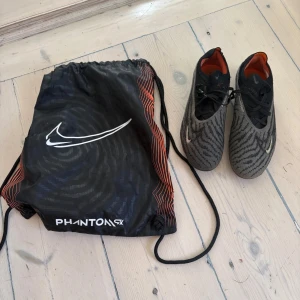 Nike Phantom GT2 fotbollsskor svart/grå - Nike Phantom GT2 fotbollsskor i svart och grått med mönstrad ovandel och orange insida. Skorna har snörning och en rund tå, tillverkade i syntetmaterial för grym känsla på planen. Säljes med original Nike PhantomSX påse.