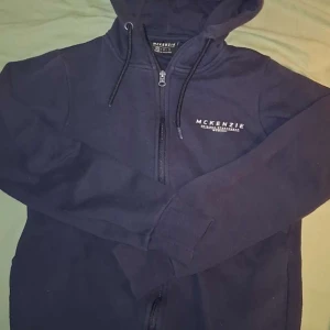 Mörkblå hoodie från McKenzie - Säljer en mörkblå hoodie från McKenzie med dragkedja och huva. Tröjan har långa ärmar, snörning i huvan och en enkel logga på bröstet. Perfekt för chill dagar eller när du vill ha en clean streetstyle-look.