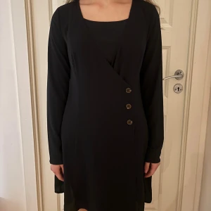 Svart omlottklänning från H&M Divided - Svart omlottklänning från H&M Divided med lång ärm och tre dekorativa knappar framtill. Storlek 40 