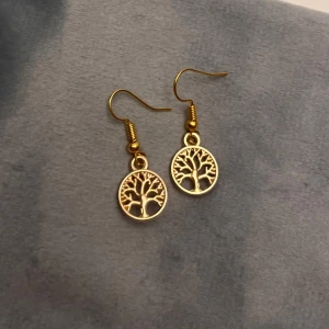 Örhängen Livets Träd Guld imitation  - Fina örhängen i form av livets träd. Berlockerna är ca 1 cm i diameter och gjorda av plast, men ser ut som metall.