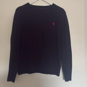 Svart kabbelstickad tröja från Polo Ralph Lauren - Svart kabelstickad tröja från Polo Ralph Lauren med klassisk röd logga broderad på bröstet. Tröjan har rund halsringning och långa ärmar, perfekt för lager-på-lager under kyliga dagar.