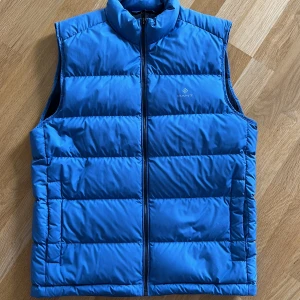 Blå dunväst från GANT, storlek M - Snygg blå dunväst från GANT i storlek M. Västen har quiltad design med horisontella sömmar, hög krage och dragkedja framtill. Perfekt att slänga över en hoodie för extra värme. Diskret GANT-logga på bröstet. Materialet är polyester och dun. Väldigt bra sick, som ny. 