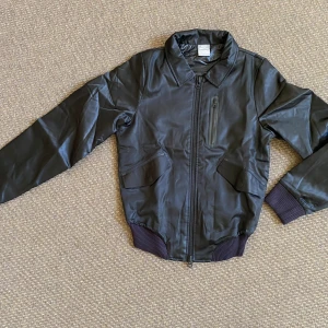 Svart bomberjacka från Nike - Snygg svart bomberjacka från Nike med klassisk krage, ribbade muddar och dragkedja framtill. Jackan har en bröstficka med dragkedja och två snedställda fickor. Materialet är glansigt och känns sportigt, perfekt för dig som gillar streetwear.