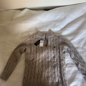 Beige stickad tröja Polo Ralph Lauren - Kabelstickad beige tröja från Polo Ralph Lauren med halv dragkedja och ribbade muddar. Klassisk design med broderad logga på bröstet och hög krage. Perfekt för lager-på-lager och snygg till jeans eller kjol. Helt ny med prislapp