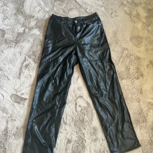 Svarta byxor i skinnimitation - Snygga svarta byxor i skinnimitation med klassisk femficksdesign och raka ben. Byxorna har knapp och dragkedja framtill och ger en cool look med sin blanka finish. Perfekta för dig som vill sticka ut med en edgy stil.