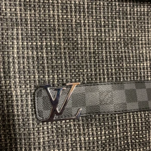 Louis Vuitton svart rutigt skärp - Säljer ett svart skärp från Louis Vuitton med klassiskt rutigt Damier-mönster och silverfärgat LV-spänne. Bältet är gjort i äkta skinn och har en stilren, exklusiv känsla. Perfekt accessoar för att lyfta din outfit med en lyxig touch.