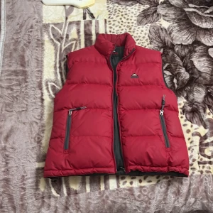 Röd dunväst från McKinley - Röd dunväst från McKinley med hög krage och två dragkedjefickor framtill. Västen har svarta detaljer och en broderad logga på bröstet. Insidan är svart med en extra ficka med dragkedja. Perfekt för lager-på-lager under kyliga dagar.