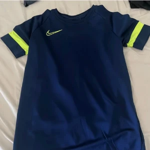 Blå Nike t-shirt med neongula detaljer - Snygg marinblå t-shirt från Nike med korta ärmar och neongula detaljer på ärmarna samt Nike-logga på bröstet. Perfekt för dig som gillar sportig stil och vill sticka ut lite extra.
