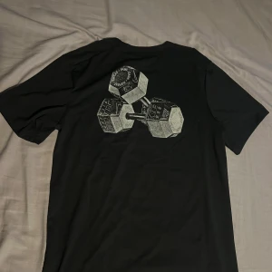 Svart Nike Dri-Fit t-shirt med tryck - Svart Nike Dri-Fit t-shirt i sportig modell med korta ärmar. Framsidan har en liten Nike-logga och baksidan är dekorerad med ett coolt tryck av två hantlar. Tillverkad i funktionsmaterial som andas och passar perfekt till gymmet.