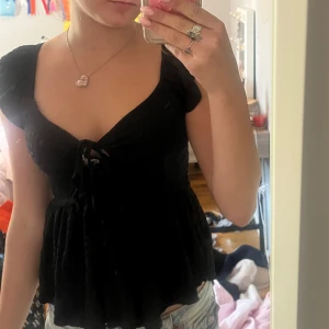 Svart blus med snörning från 157 - Supersöt svart blus från 157 i storlek XS. Modellen har korta ärmar, v-ringning och snörning framtill. Tyget är lätt och har en lite rynkad struktur som ger en flowy känsla. Perfekt att styla med jeans eller kjol för en trendig look.
