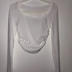 Vit ribbad långärmad topp - Säljer en vit långärmad topp med ribbat tyg och diskreta vertikala ränder. Toppen har en rundad halsringning och snygg rynkdetalj i sidorna som ger en cool form. Perfekt att styla med jeans eller kjol för en fräsch look.
