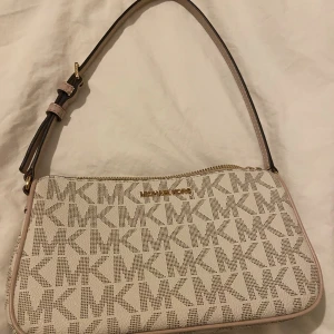Beige axelväska från Michael Kors - Snygg beige axelväska från Michael Kors med justerbart axelband och guldiga detaljer. Väskan har MK-logga över hela ytan och stängs med dragkedja. Perfekt storlek för det viktigaste och ger en clean look till din outfit.