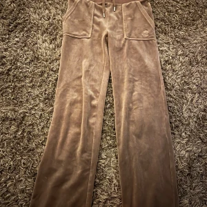 Beige mjukisbyxor i velour Juicy Couture - Supermjuka beige mjukisbyxor från Juicy Couture i velour med raka ben, stora fickor fram och bak samt snörning i midjan. Perfekta för en chill och avslappnad look. Klassisk logga broderad på ena fickan framtill. Använda fåtal gånger