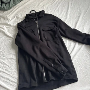 Svart overshirt med dragkedja - Svart overshirt från H&M med två stora bröstfickor och hel dragkedja framtill. Jackan har klassisk krage, långa ärmar med knappdetalj vid ärmslut och är tillverkad i mjukt material. Perfekt för lager-på-lager och enkel att styla.