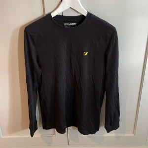 Svart långärmad tröja från Lyle & Scott - Svart långärmad tröja från Lyle & Scott med klassisk rund hals och gul broderad logga på bröstet. Tillverkad i mjuk bomull som är skön mot huden. Perfekt för dig som gillar stilrena och enkla plagg med en sportig touch.