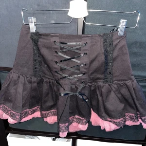 Svart kortkjol med rosa spets från Hell Bunny - Supersöt svart kortkjol från Hell Bunny med snörning framtill, volangkant och rosa spetsdetaljer längst ner. Kjolen har dragkedja bak och coola svarta spetsband på sidorna. Perfekt för dig som gillar alternativ stil och vill sticka ut.