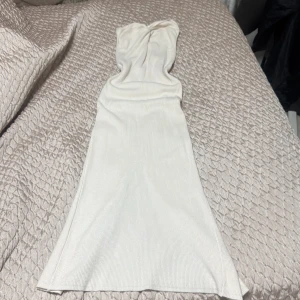 Vit axelbandslös långklänning - Säljer en vit axelbandslös långklänning i ribbat material. Klänningen har en twistad detalj vid bysten och en snygg slits bak för extra rörelsefrihet. Perfekt för dig som gillar stilrena och eleganta plagg med en modern touch.