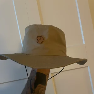 Beige Fjällräven hatt  - Snygg beige hatt från Fjällräven med bred brätte och klassisk rävlogga framtill. På sidan finns en liten svensk flagga. Hatten har justerbar snodd under hakan och är tillverkad i slitstark polyester, perfekt för soliga dagar.