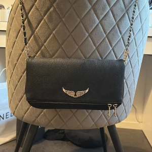 Svart axelväska med kedjerem - Snygg svart axelväska i skinnimitation med silverfärgad kedjerem och detaljer. Kommer med en längre axelrem och en kortare, axelremmen har trasslat sig lite men om man har tålamod går det att fixa! Säljer pga att den inte kommer till användning. Väskan är inte äkta!