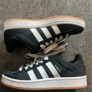 Adidas Campus sneakers svart/vit - Svarta Adidas Campus sneakers i mocka med vita klassiska ränder och vit sula. Snygg retrostil med låg profil och platt gummisula. CAMPUS-tryck på sidan och vita skosnören. Perfekta för dig som gillar streetwear och tidlös design.