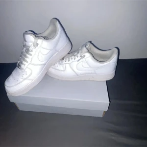 Nike Air Force 1 helvita sneakers - Klassiska Nike Air Force 1 sneakers i helvitt skinn med rund tå och platt sula. Snygg och tidlös modell med snörning och diskret Nike-logga på sidan. Perfekta för dig som vill ha en clean och stilren look till vardags. Levereras i originalkartong.