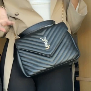 Svart quiltad axelväska från YSL - Säljer en svart quiltad axelväska YSL med guldiga detaljer och kedjerem. Väskan har ett stilrent V-format mönster och klassisk YSL-logga framtill. Tillverkad i skinn och har både handtag och axelrem för flexibel användning.