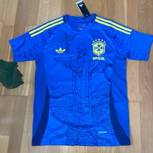 Blå Brasil Jesus Adidas fotbollströja S - Snygg blå Brasilien fotbollströja från Adidas med gula detaljer och tre ränder på axlarna. Framsidan har tryck av Kristusstatyn och CBF-emblem. Slim fit-modell.