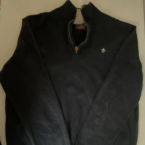 Mörkblå half zip tröja från Morris - Mörkblå långärmad tröja från Morris med half zip och ribbad krage i kontrastfärg. Tröjan har en liten broderad logga på bröstet och en patch nertill. Perfekt för lager-på-lager och snygg till jeans. Tröjan är i storlek M men sitter som S