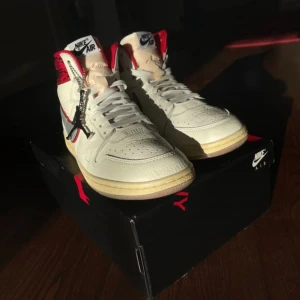 Jordan nike Air Jordan 1 Awake NY Highs - Nike Air Jordan 1 Awake NY i vitt skinn med grå swoosh och röda detaljer. Hög modell med röd och svart ormskinnsmönstrad häl, Awake NY-logga och klassisk Jordan hangtag. Snygg kontrast mellan den ljusa ovandelen och den beige sulan. Perfekta för dig som vill sticka ut.