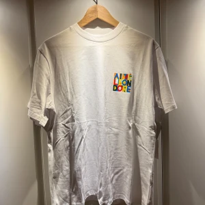 Vit t-shirt från Aimé Leon Dore - Vit t-shirt från Aimé Leon Dore med färgglad grafisk logga på bröstet och ryggen. Klassisk passform med rund hals och korta ärmar. Tillverkad i mjuk bomull, perfekt för en clean och stilren look.