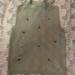 Grönt linne med blommor - Supersött linne i ljusgrön nyans med broderade små blommor i rosa och blått. Linnet har ribbad hög krage och ett diskret hålmönster i tyget. Perfekt att styla över annat plagg för en trendig lager-på-lager-look.