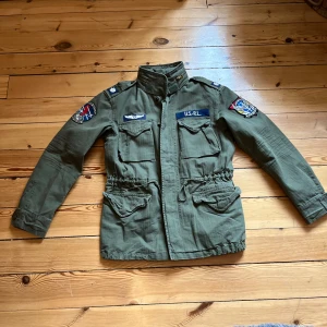 Olivgrön parkajacka Polo Ralph Lauren - Snygg olivgrön parkajacka från Polo Ralph Lauren med coola patchar och broderade detaljer på ärmarna. Jackan har hög krage, fyra stora fickor framtill och dragsko i midjan för en schysst passform. Om du har några frågor så tveka inte på att skriva eller om du vill ha fler bilder! Ny pris på den här jackan är 5000kr. Den är i jätte bra sick nästan aldrig använd! Passar dig runt 170!